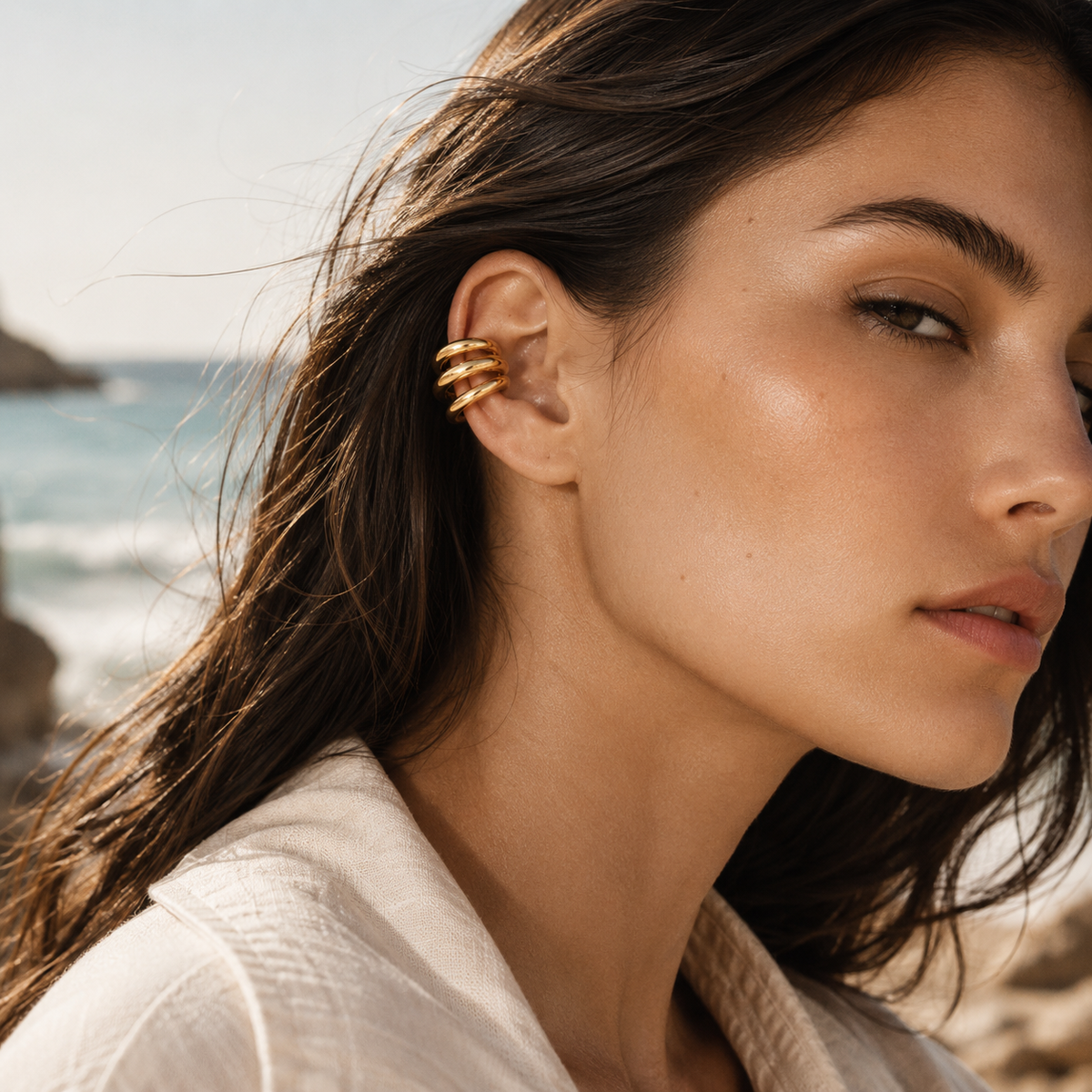 The Dréa EAR CUFF worn on the ear at the beach, a multi-band gold cuff on a model in a linen jacket.