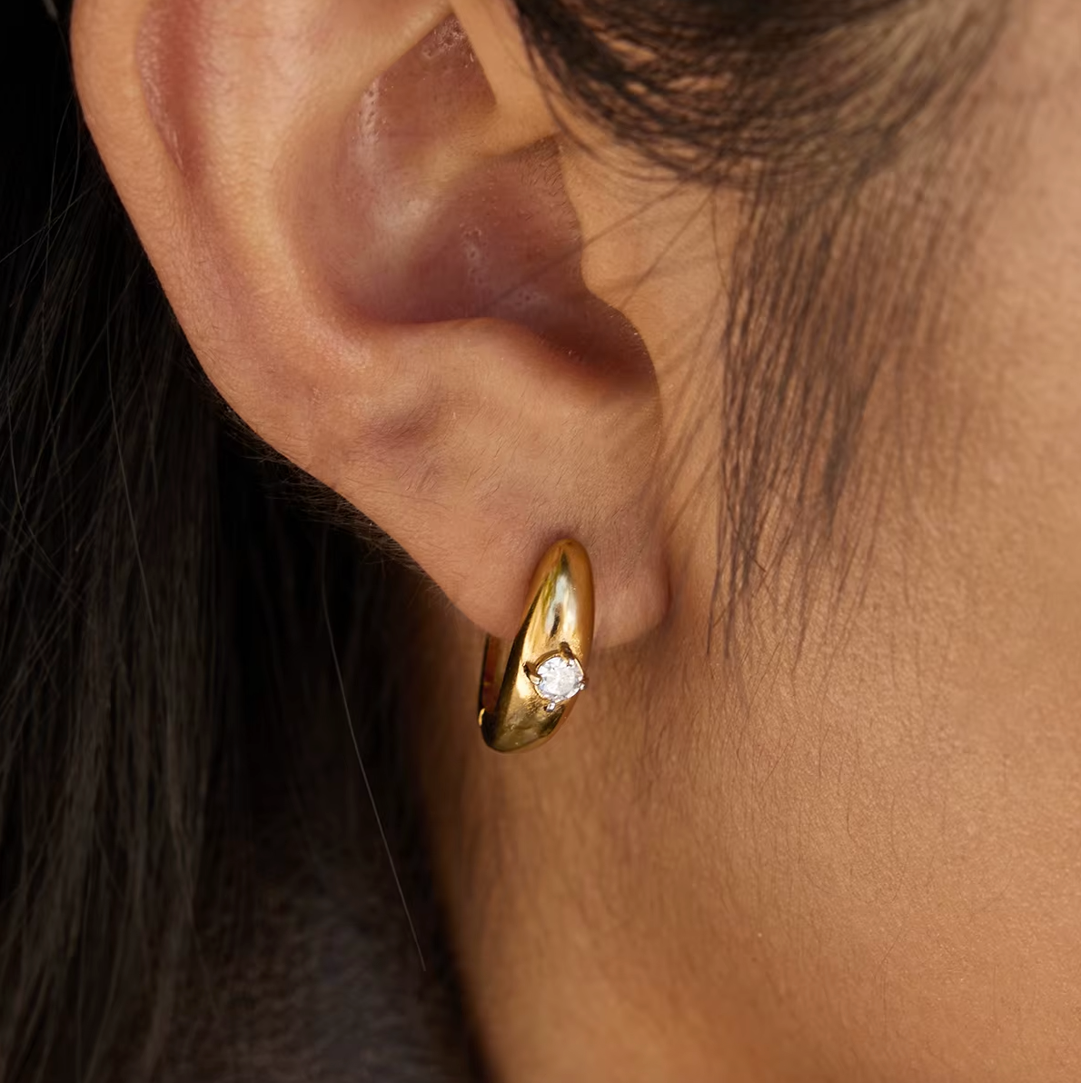 Worn close on a model's ear, the Everyday Drama EARRINGS show their smooth gold curve and single crystal.