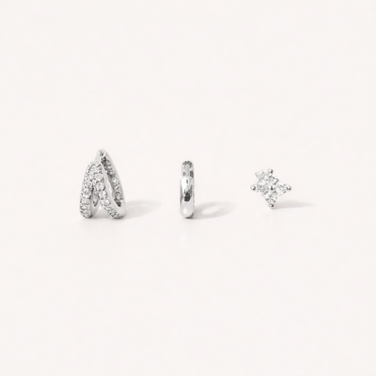 Faye Set in silver tone, a pave crossover hoop, a plain huggie hoop, and a four-point crystal stud, laid flat.