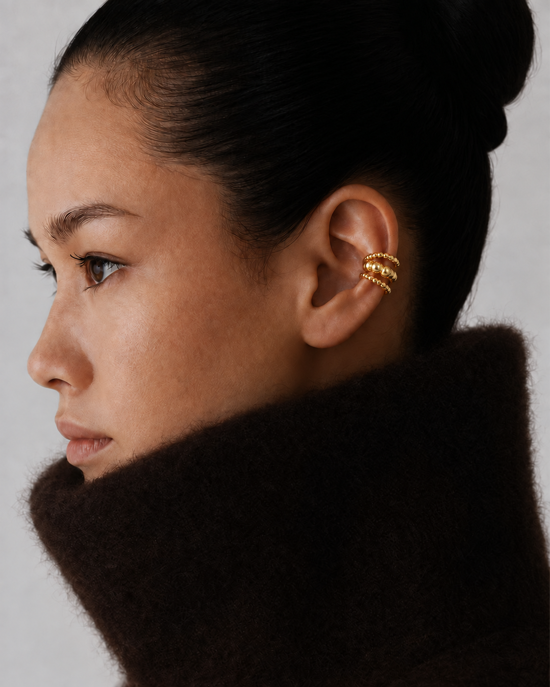 Close-up of a model's ear wearing the gold Folie à Trois Felix EAR CUFF's triple beaded band, styled with a chunky knit collar.