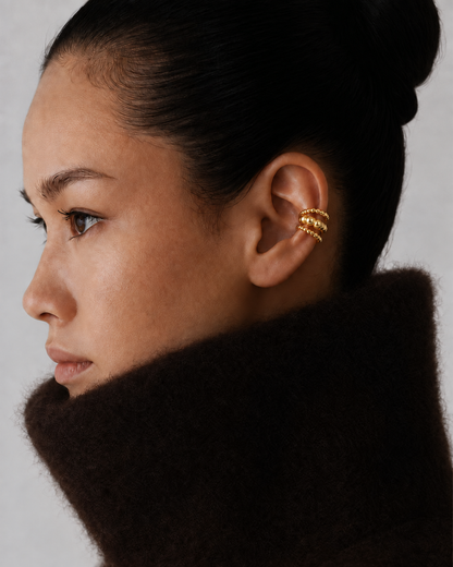 Close-up of a model's ear wearing the gold Folie à Trois Felix EAR CUFF's triple beaded band, styled with a chunky knit collar.