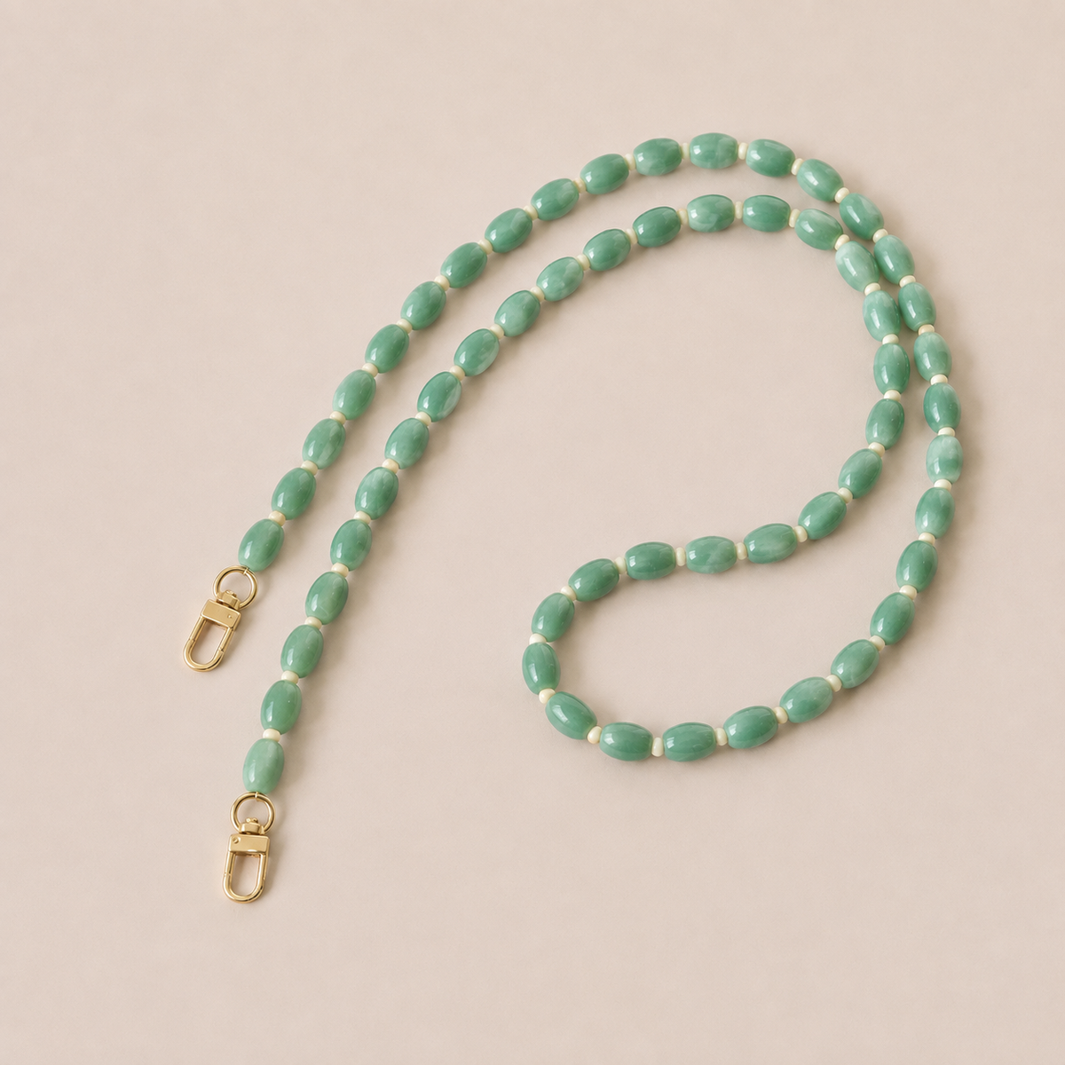Folie à Trois Fennel long wholesale phone chain in jade green oval beads with cream spacers and gold clasps, laid flat on a cream background.
