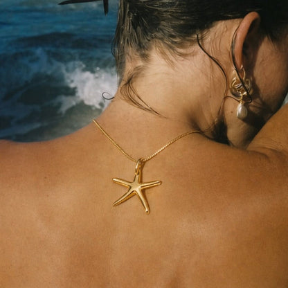 The Fiji NECKLACE worn from behind at the nape of the neck, its gold starfish resting against tanned skin with the ocean behind.