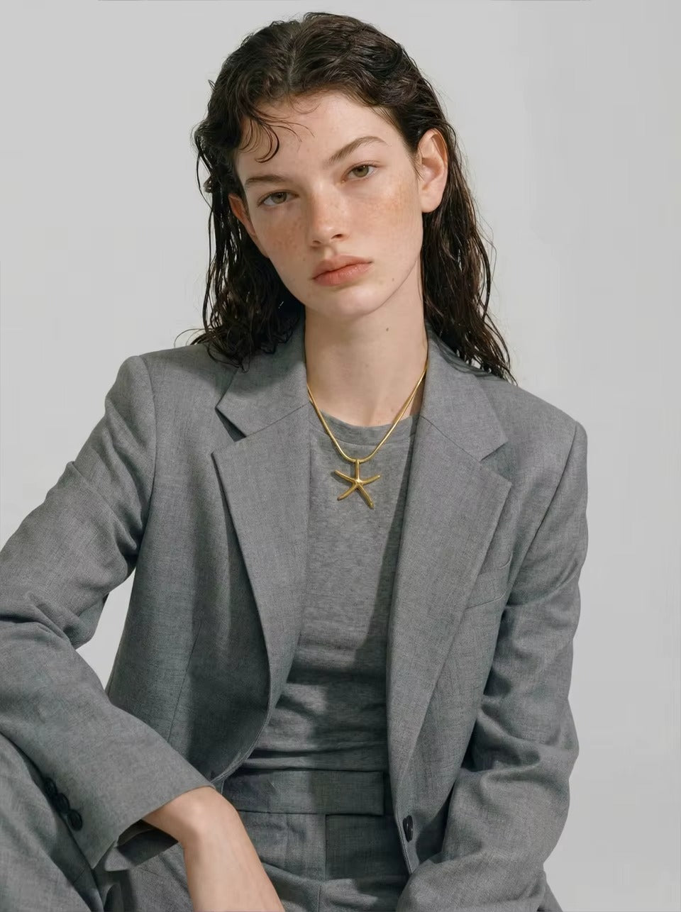 The Fiji NECKLACE worn layered with a thin chain over a gray tee and blazer, the model seated with her arm resting forward.