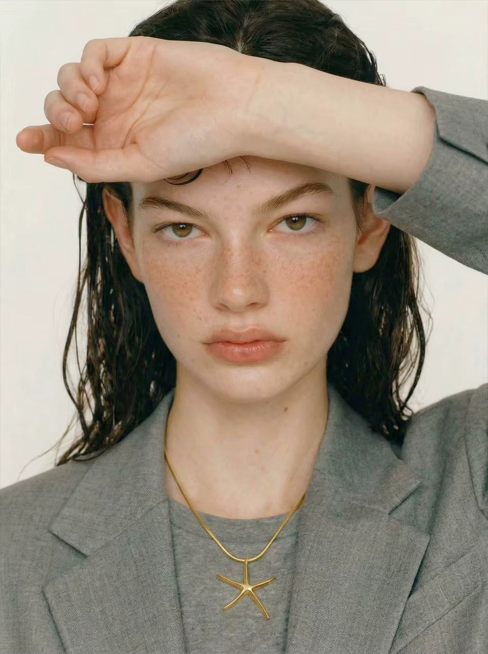 The Fiji NECKLACE worn close-up on a gray tee, the model shielding her eyes with wet hair falling over her shoulders.