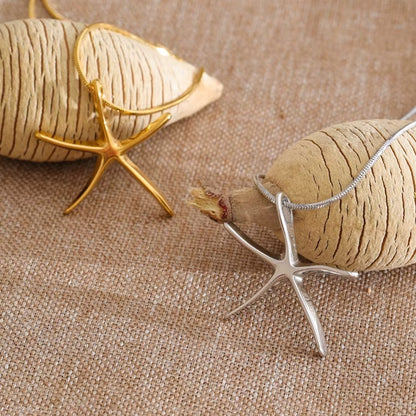 The Fiji NECKLACE shown in gold and silver from a different angle, both starfish charms resting against woven fabric and shells.