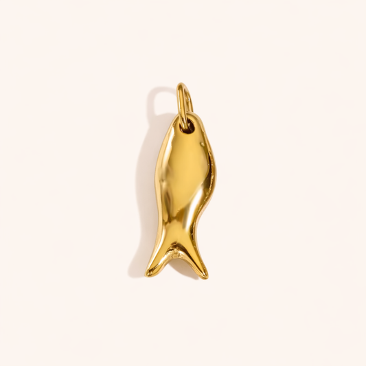 The Fish CHARM in a studio flat lay, a sculptural polished gold pendant shaped like a swimming fish on a cream background.