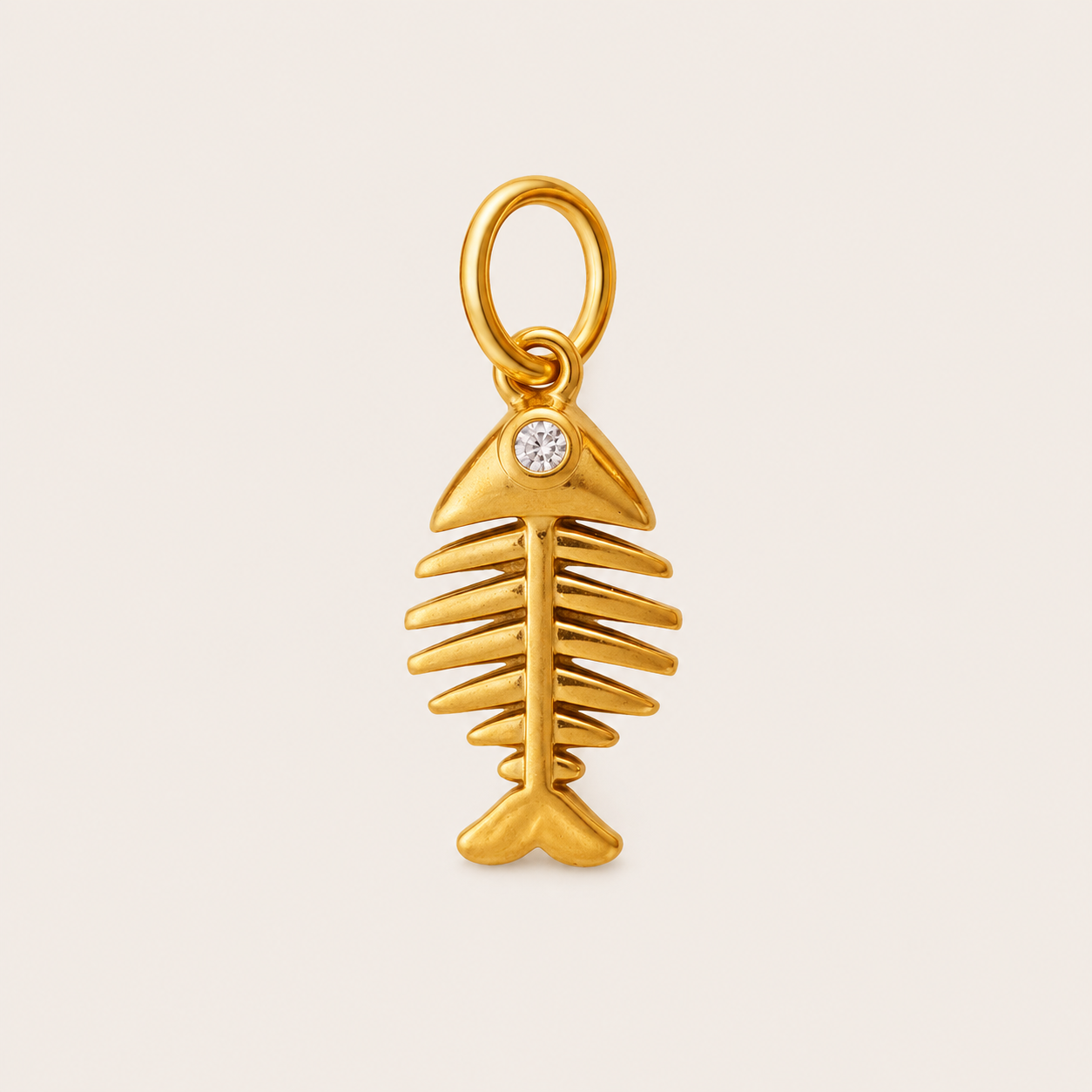 A gold fish skeleton charm pendant with a small clear round gemstone set in the gill area, shown on a light beige background.