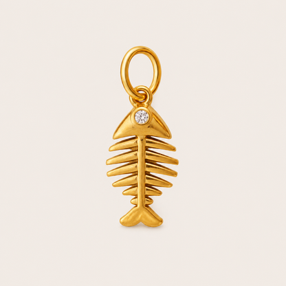A gold fish skeleton charm pendant with a small clear round gemstone set in the gill area, shown on a light beige background.