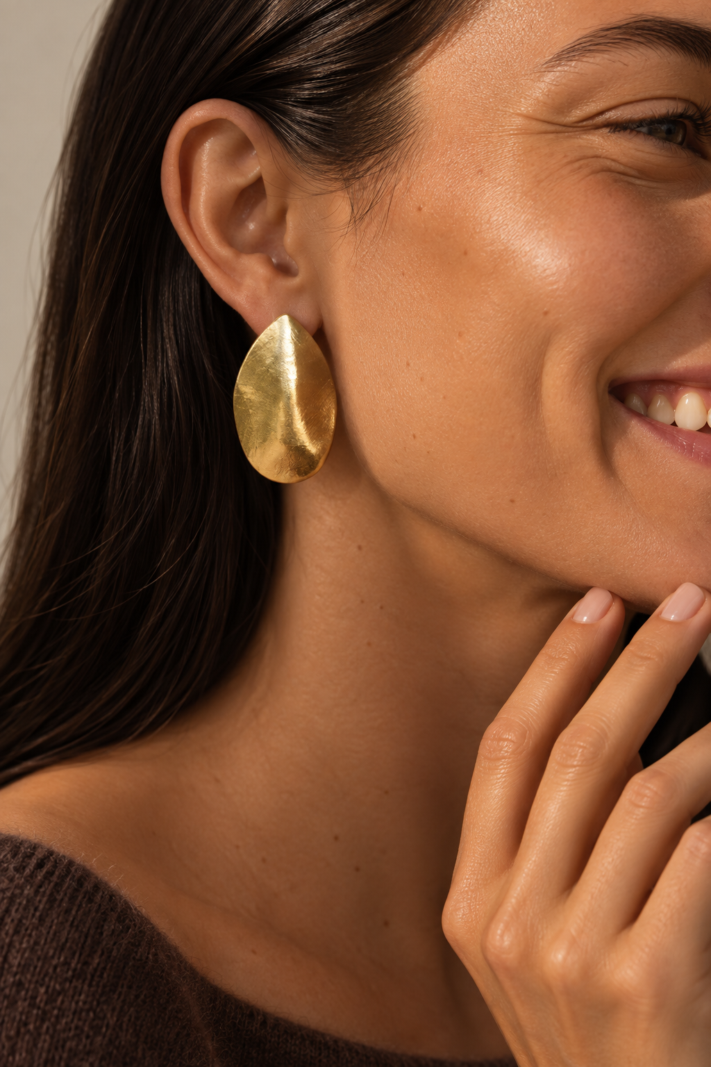 Model wears a large gold leaf-shaped Foglia EARRINGS stud, smiling with hand near her chin — Folie à Trois
