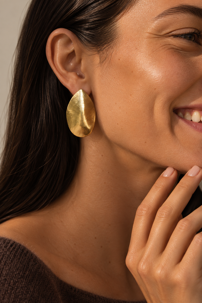Model wears a large gold leaf-shaped Foglia EARRINGS stud, smiling with hand near her chin — Folie à Trois