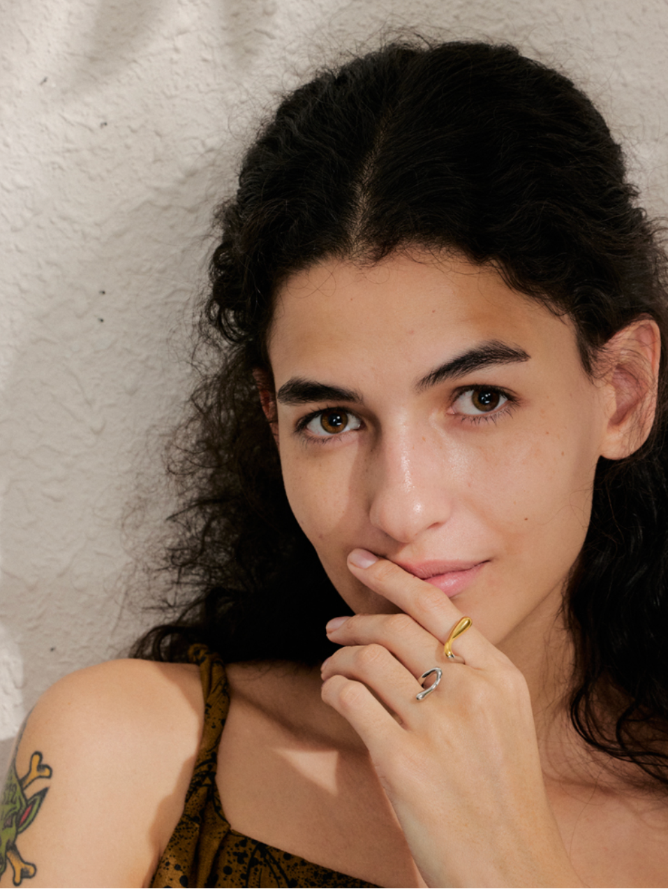 Close-up of a woman's face and hand wearing the gold Frances RING stacked with a silver hook-shaped ring, near her chin.