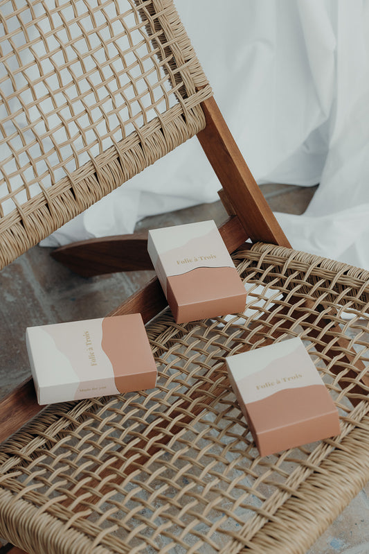Three rust and cream Folie à Trois gift boxes resting on a woven rattan chair against a white curtain backdrop.