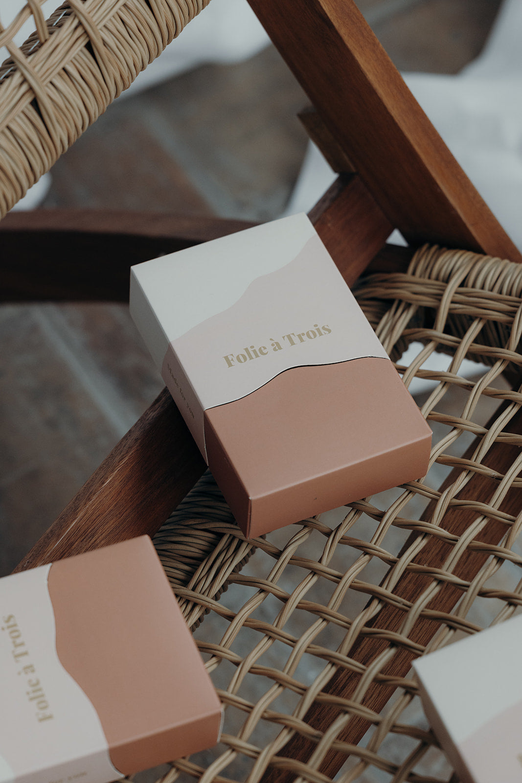 Close-up of a rust and cream Folie à Trois gift box angled on a rattan chair beside two more boxes.