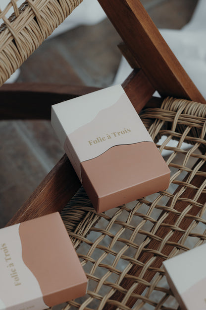 Close-up of a rust and cream Folie à Trois gift box angled on a rattan chair beside two more boxes.