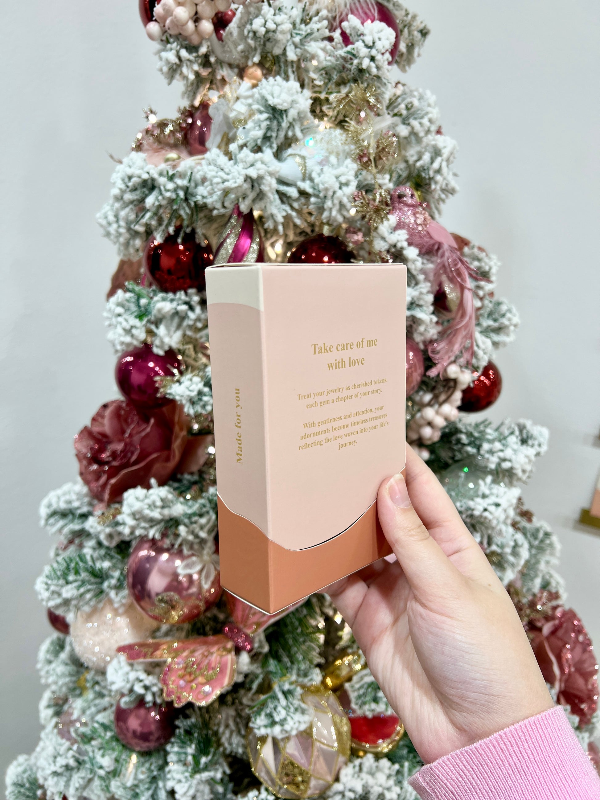 Hand holding an open Folie à Trois gift box displaying its care message against a festive Christmas tree backdrop.