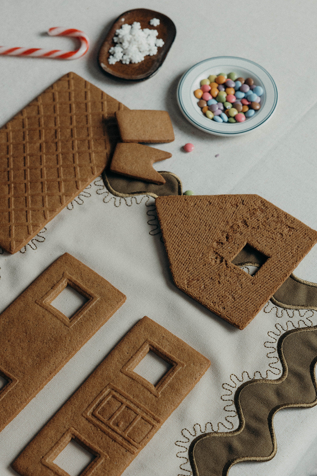 Gingerbread house cookie pieces and candy displayed beside the olive Gisèle Scallop placemat's gold-stitched scalloped edge.