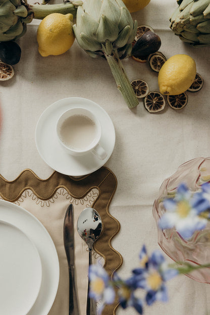 Overhead table setting with a white cup, plates, and blue flowers arranged on the olive Gisèle Scallop placemat with embroidered trim.