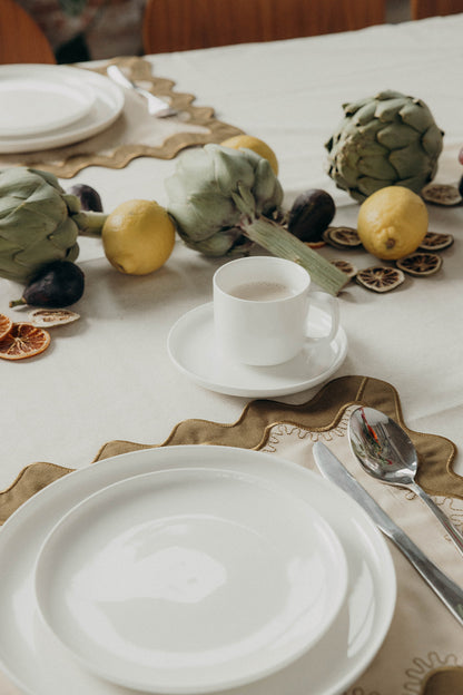 Table setting with white plates, cutlery, and a coffee cup arranged on the olive Gisèle Scallop placemat with a fruit garland nearby.