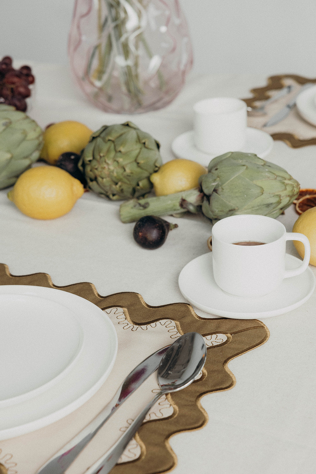 Close angle of a white plate, cutlery, and coffee cup on the olive Gisèle Scallop placemat with lemons and artichokes in the background.