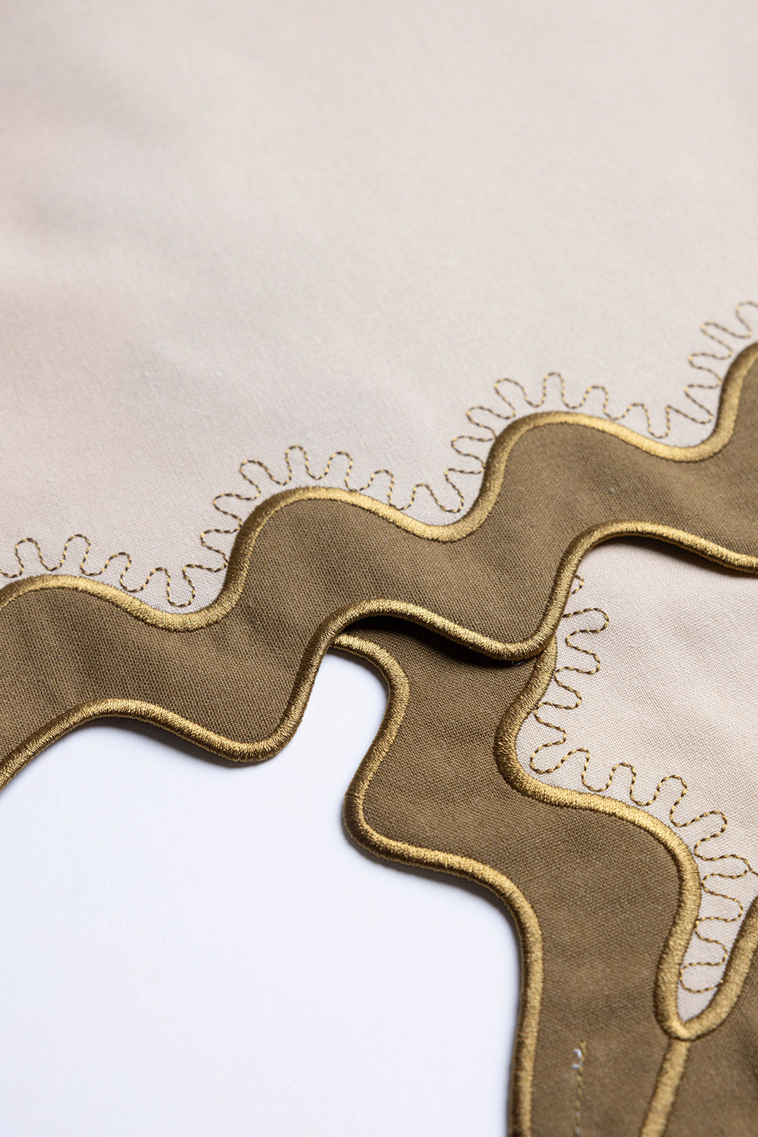 Macro shot of a gold-stitched scalloped edge on cream and olive fabric, detail of the Gisèle Scallop napkins with embroidered trim.