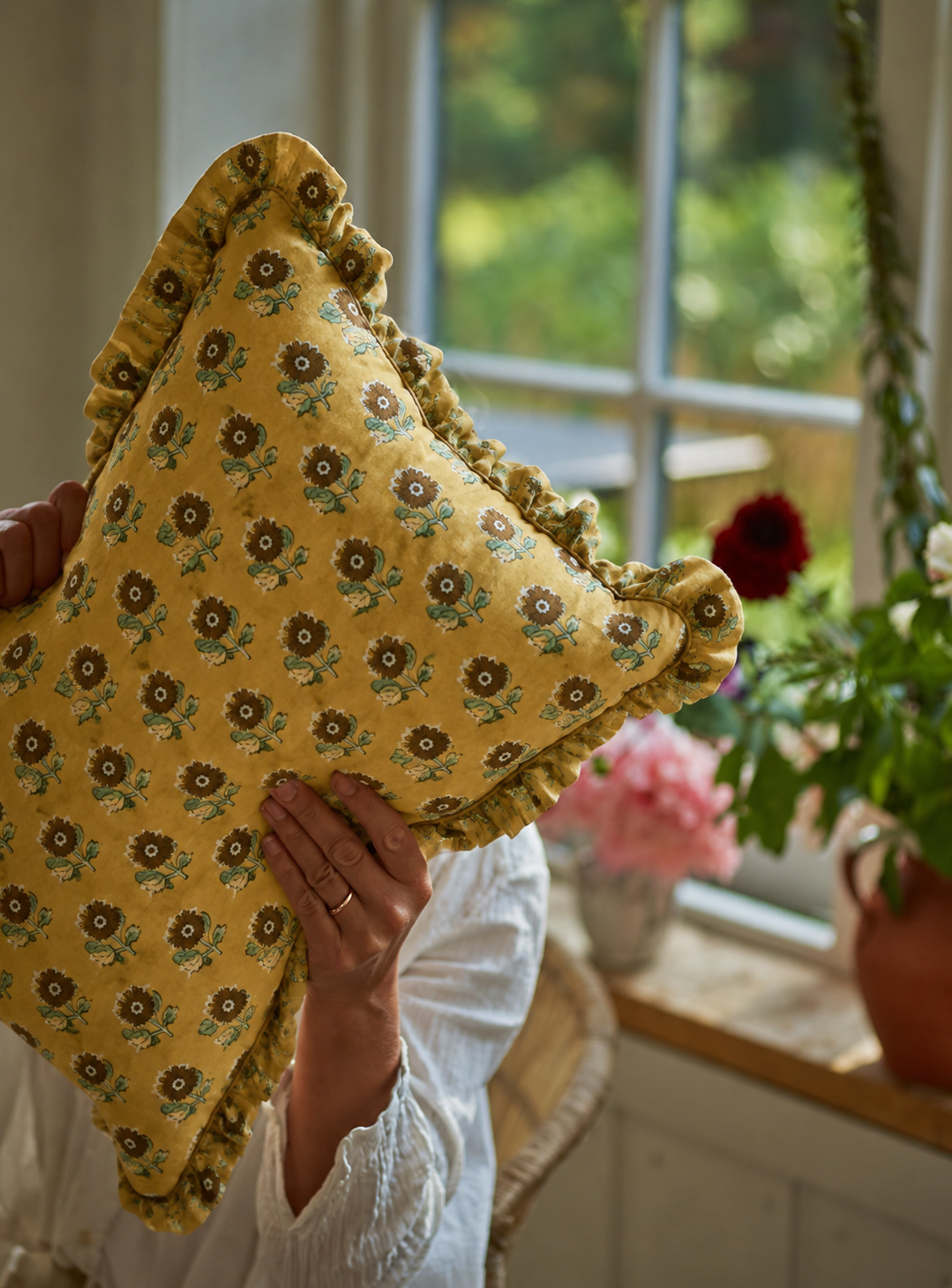 Golden Petal Rectangle cushion held up indoors near a sunlit window, its mustard sunflower print framed by fresh flowers on the sill.