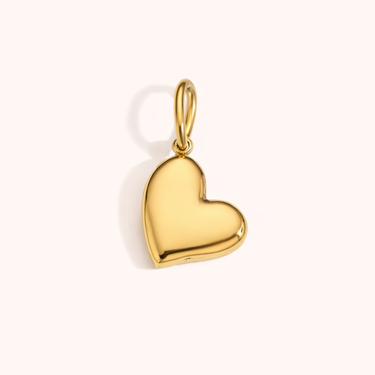 The Heart CHARM in a studio flat lay, a polished gold puffed heart pendant catching the light on a cream background.