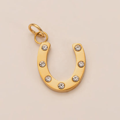 The Horseshoe CHARM in gold, a curved pendant set with clear zircon stones, shown in close-up studio photography on cream.