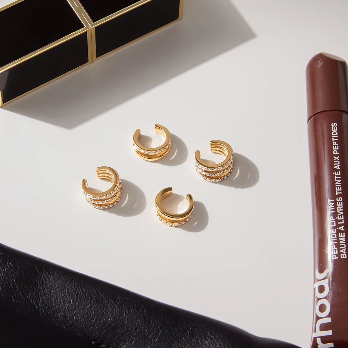 Two Hotline Ear Cuffs arranged on a white marble surface next to a lip tint tube, shown alongside a fuller pave crystal cuff pair for scale.