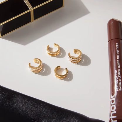 Two Hotline Ear Cuffs arranged on a white marble surface next to a lip tint tube, shown alongside a fuller pave crystal cuff pair for scale.