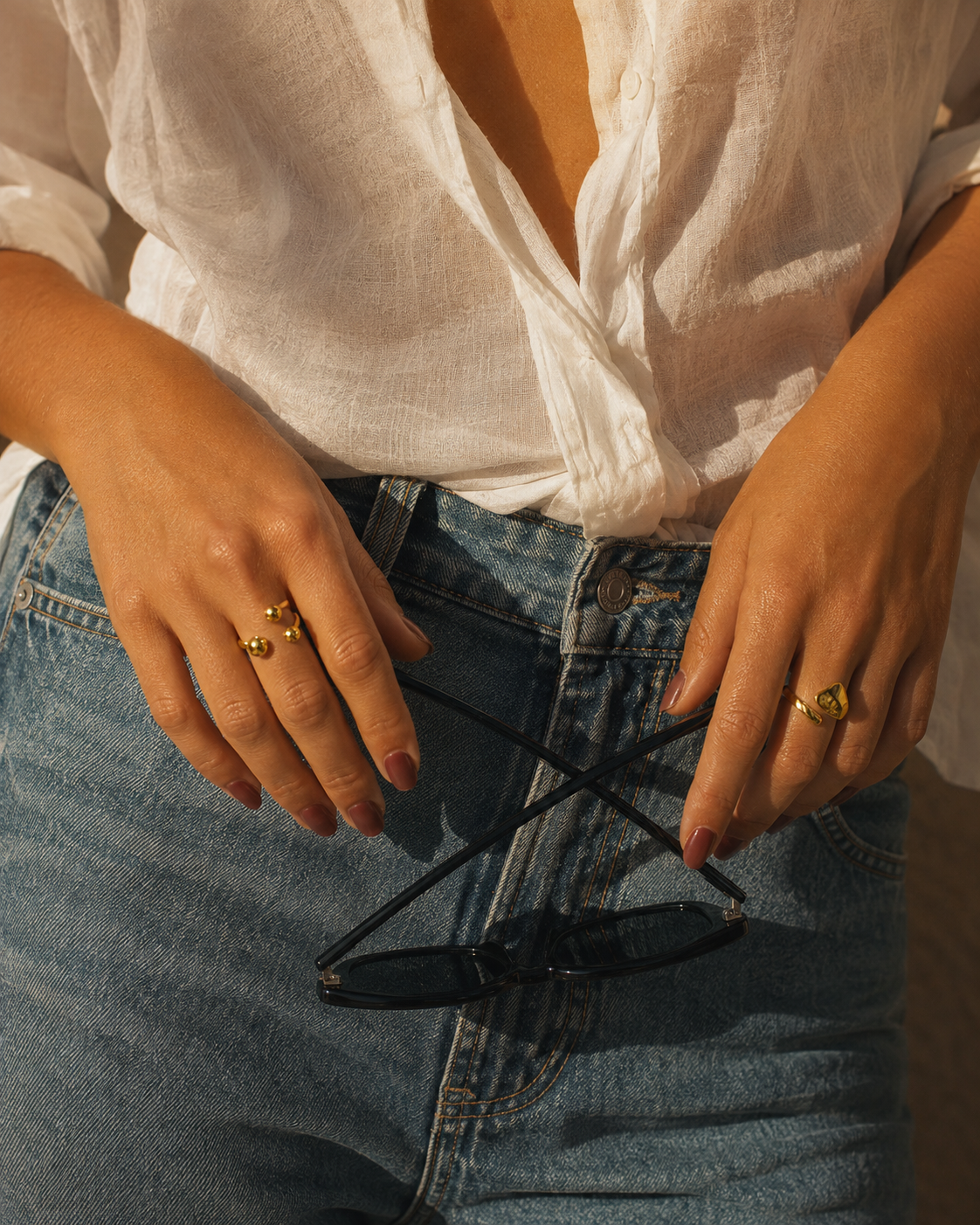 Model's hands rest near denim jeans wearing the gold Ibiza RING along with another delicate gold ring, sunglasses draped between them.