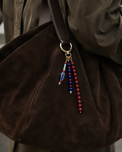 Folie à Trois Isadora BAG CHARM in navy, red and cream beaded strands, clipped to a dark brown suede shoulder bag.