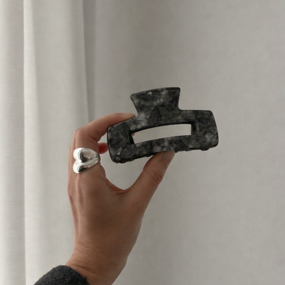 It Girl Big HAIR CLIP's charcoal grey marbled claw clip held up by hand against a curtain, styled with a silver heart ring.