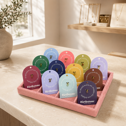 Pink velvet jewelry tray filled with colorful Birthstone charm cards on a marble counter beside a window and plant.