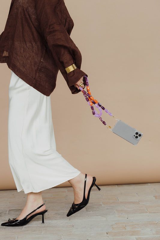 A woman walking while holding a phone by its purple and orange bead chain, wearing a brown linen top and gold cuff.