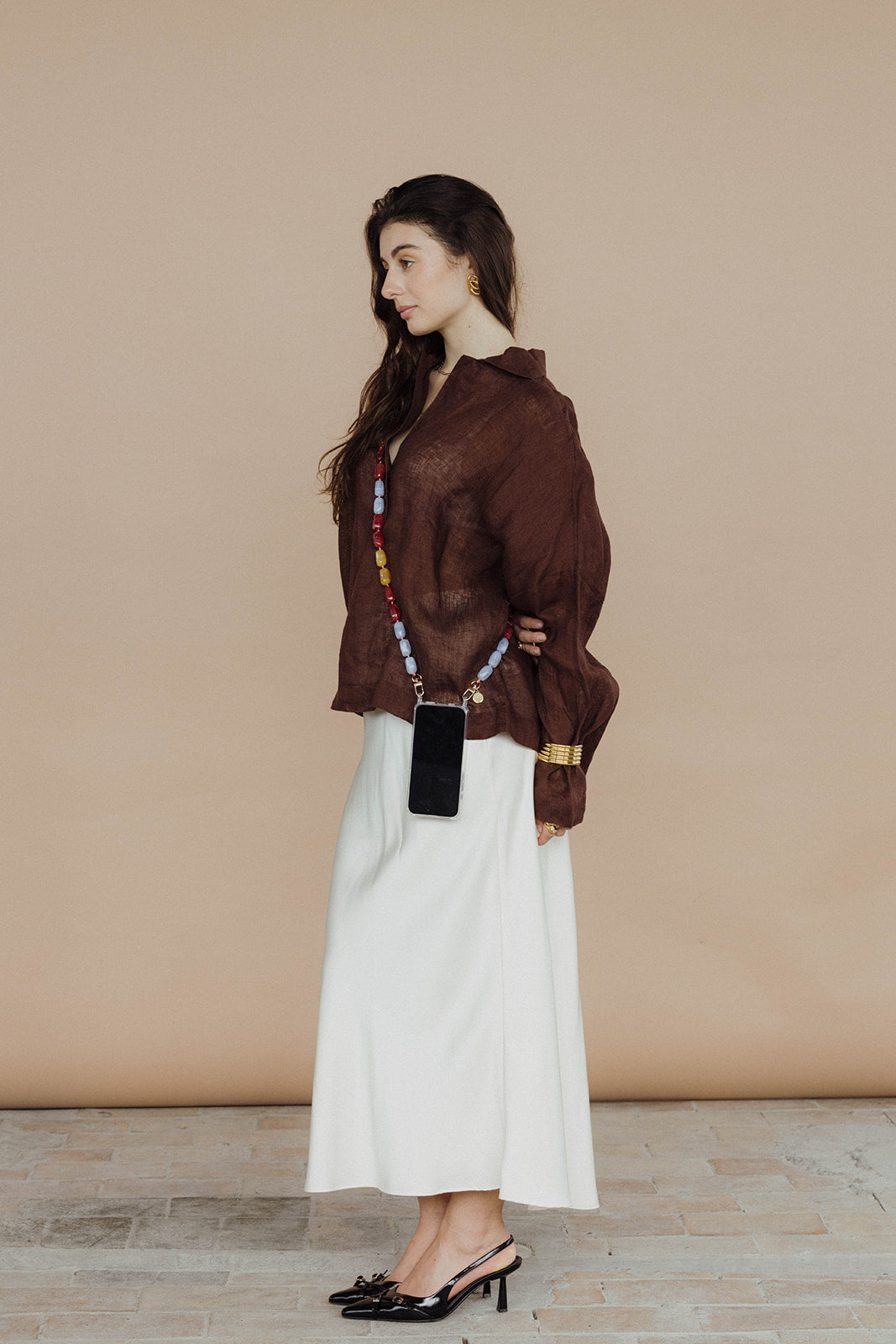 A woman in a brown linen top and white skirt, wearing a lavender, red, mustard, and steel blue bead phone chain crossbody.
