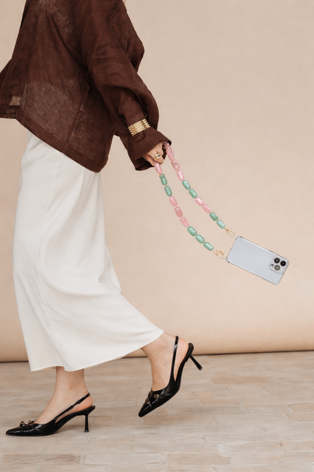 A woman walking while holding a phone by its pink and mint green bead chain, wearing a brown linen top and gold cuff.