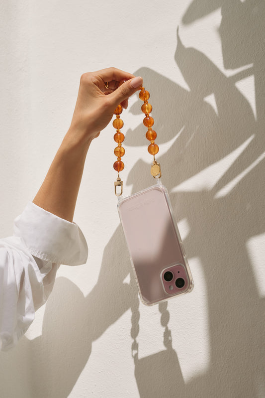 A hand holding a clear phone case by its amber bead phone strap, catching sunlight against a white wall, with a white sleeve visible.
