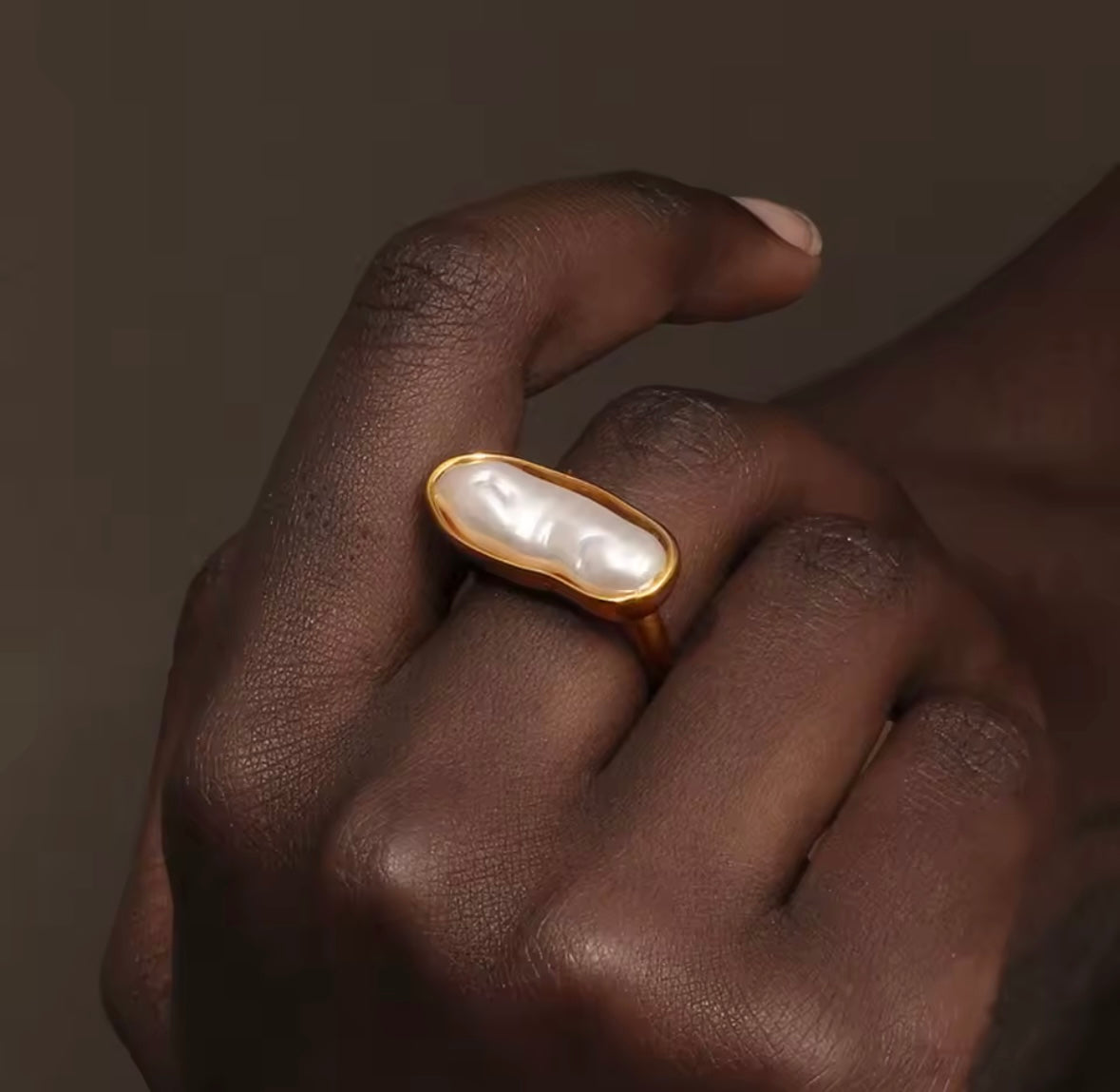 Close-up of a hand wearing the gold La Mer RING with its elongated baroque pearl, against a warm brown backdrop.