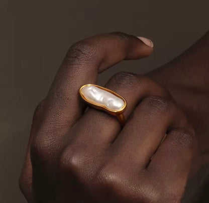 Close-up of a hand wearing the gold La Mer RING with its elongated baroque pearl, against a warm brown backdrop.