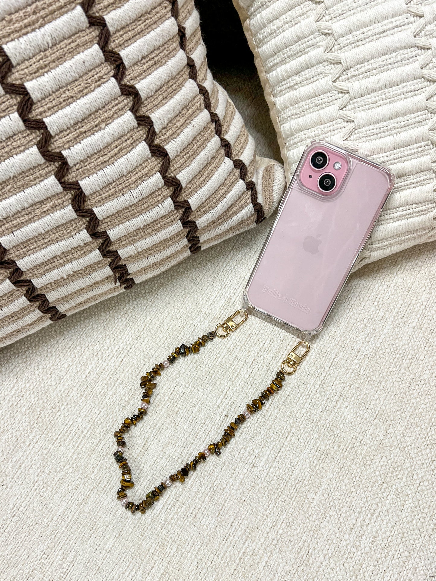 Brown and gold chip-bead Lagoon Stones SHORT PHONE STRAP attached to a pink phone case, resting on woven cushions.