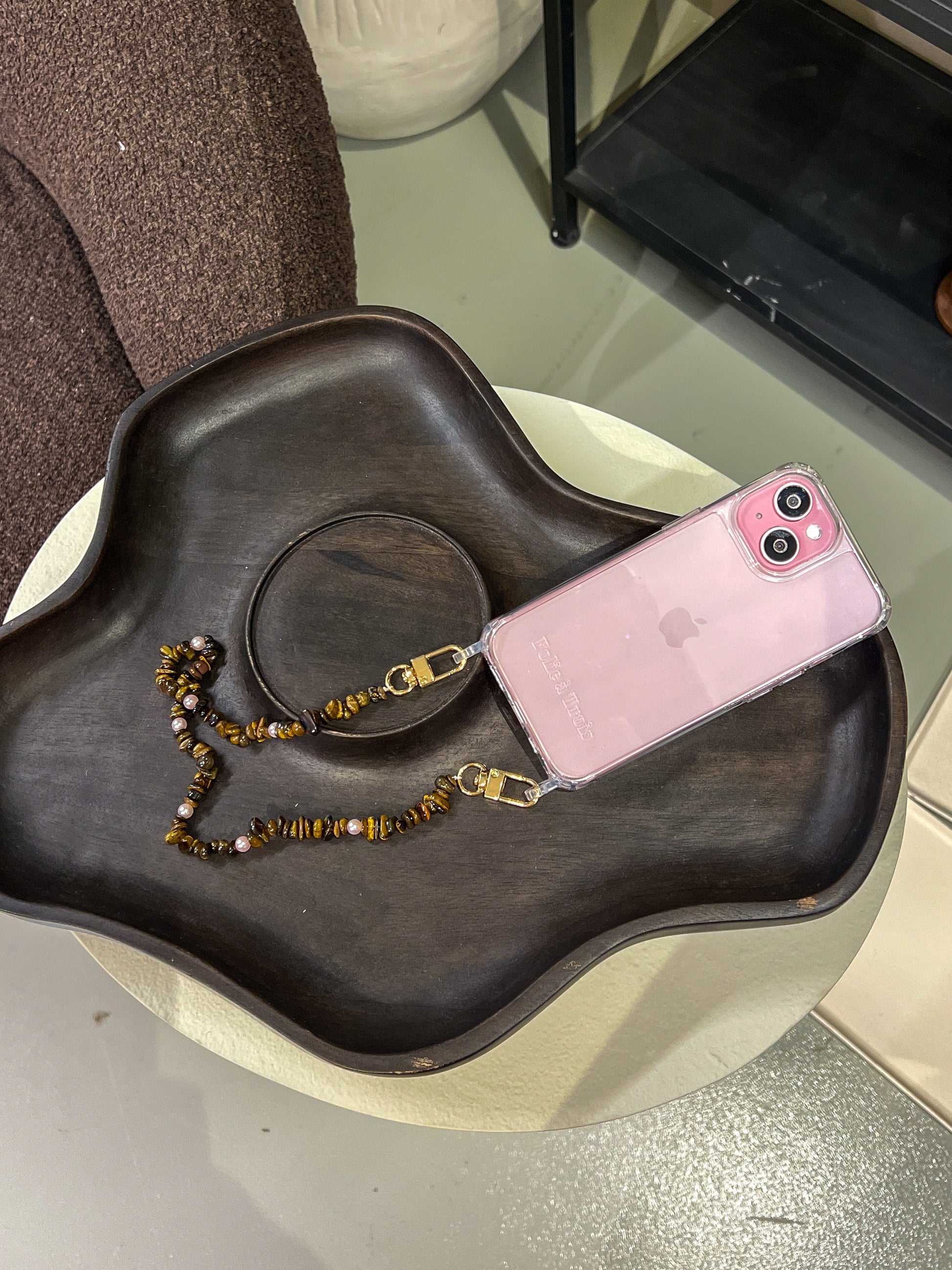 Brown and gold chip-bead Lagoon Stones SHORT PHONE STRAP attached to a pink phone case, styled on a wooden tray.