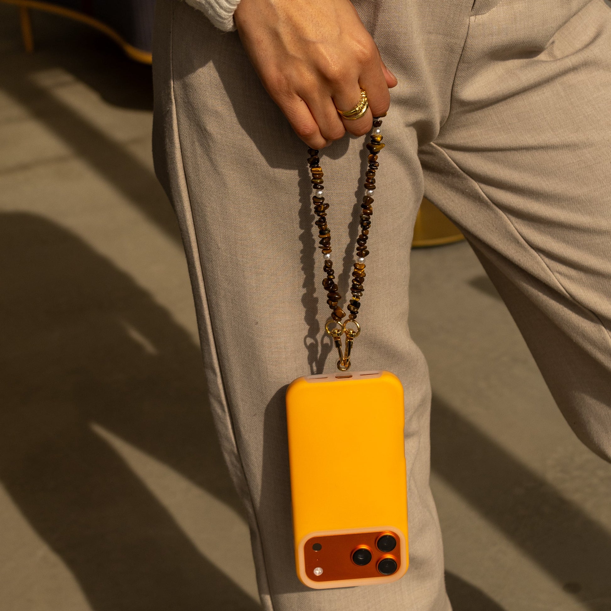 Woman holding an orange phone case attached to a brown and gold chip-bead Lagoon Stones SHORT PHONE STRAP.