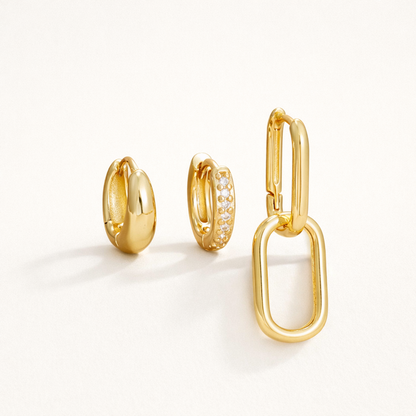 Lea Set of gold jewelry, a chunky teardrop hoop, a pave hoop, and a paperclip hoop with a large oval charm, laid flat.