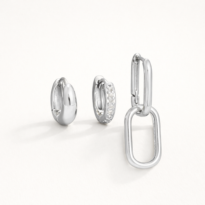 Lea Set in silver tone, a chunky teardrop hoop, a pave hoop, and a paperclip hoop with a large oval charm, laid flat.