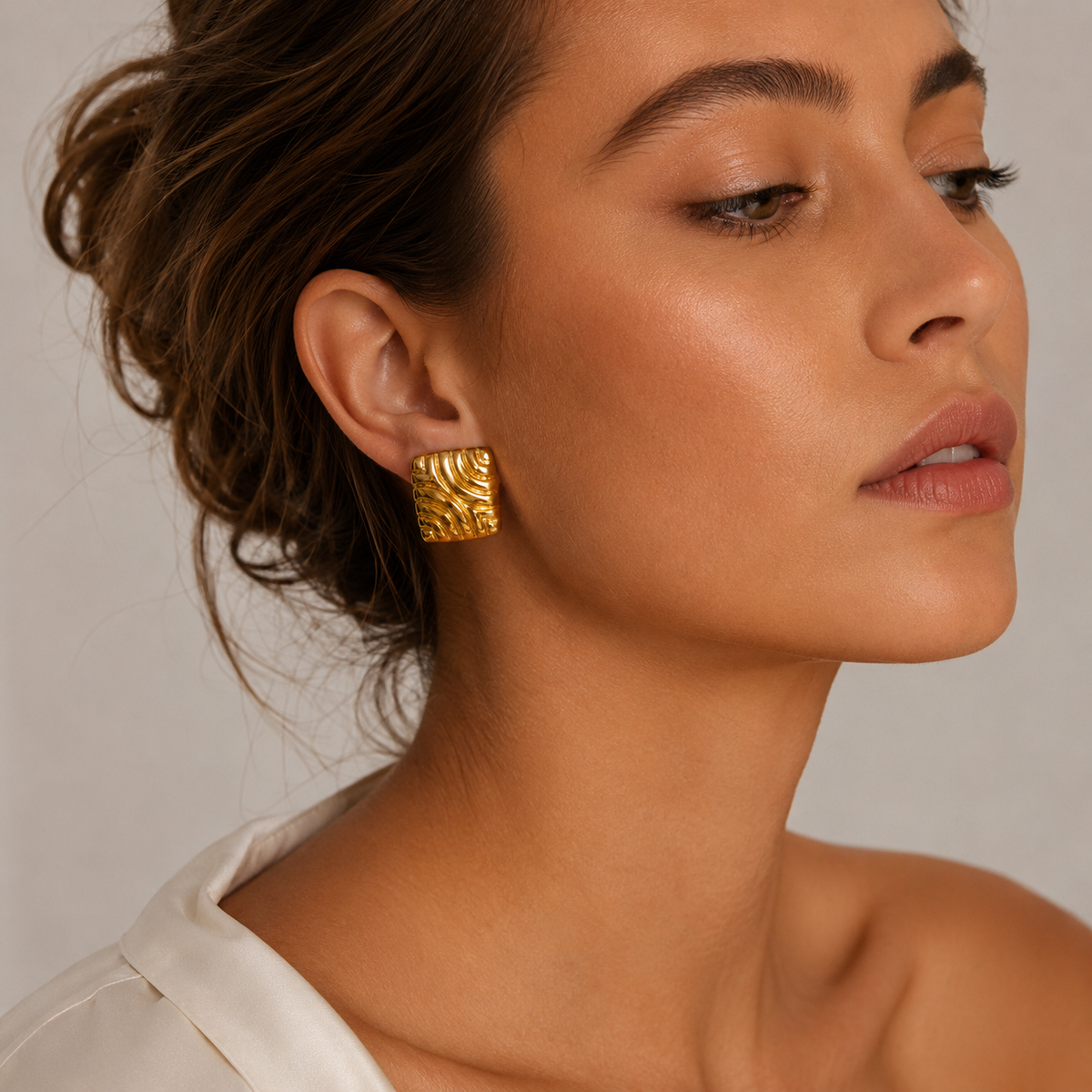 Close-up of a model wearing a gold Leia EARRINGS square stud with a swirling, ribbed wave texture and polished finish.