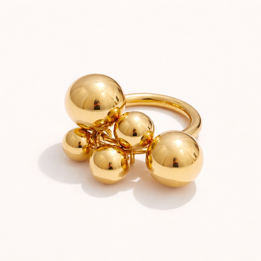 Gold ring with a cluster of five polished ball charms dangling from the band, shown at an angle on a cream backdrop — Lennox RING.