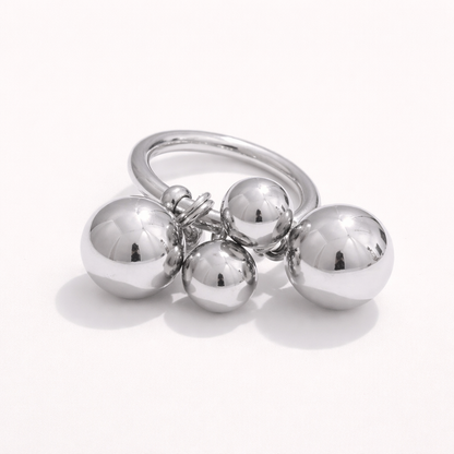 Silver ring with a cluster of four polished ball charms dangling from the band, shown at an angle on a cream backdrop.