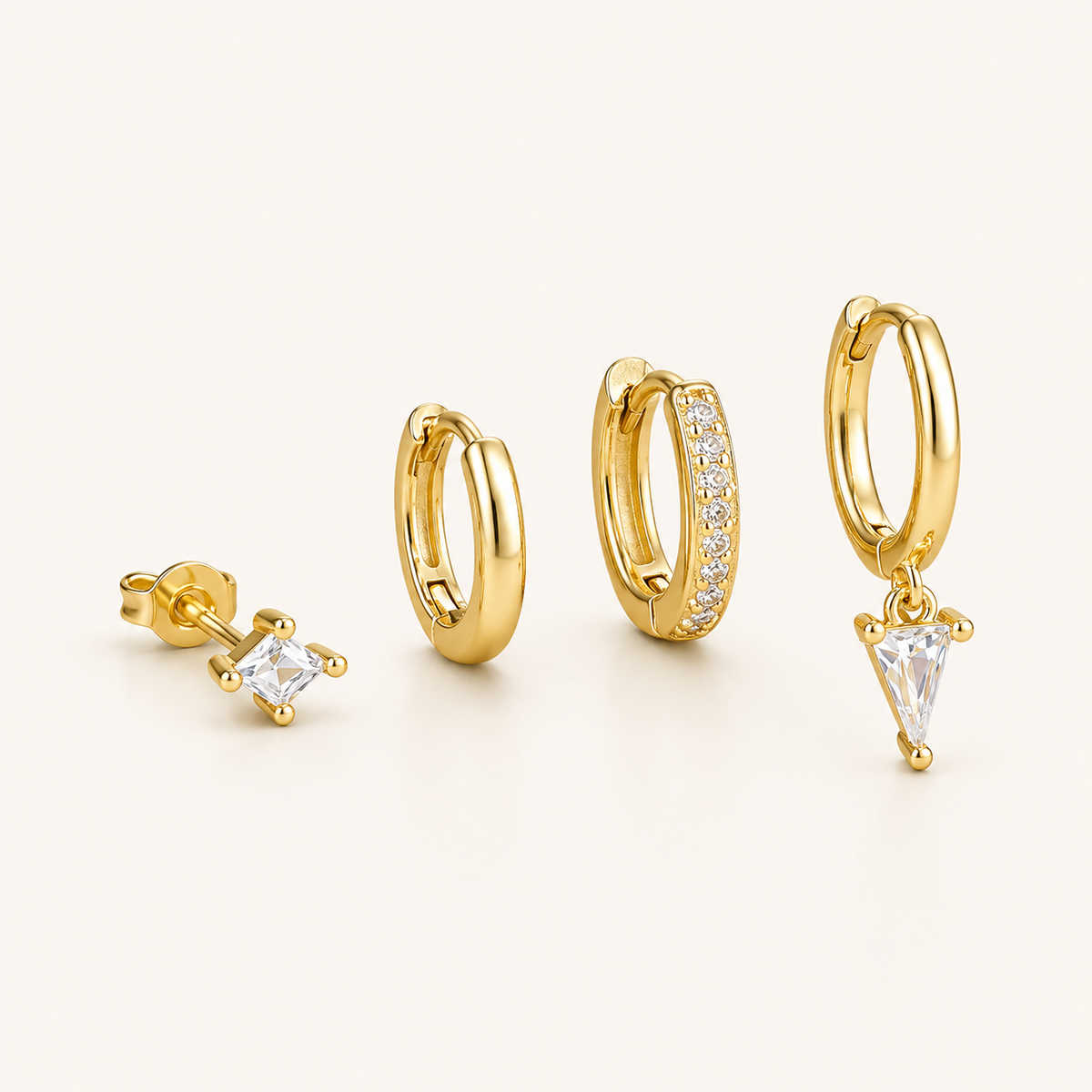 Lexi Set of gold jewelry, a princess-cut stud, a plain hoop, a pave hoop, and a hoop with a triangle crystal drop, laid flat.