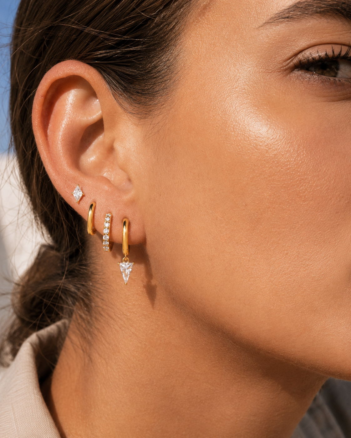 Lexi Set worn on the ear in gold, a princess-cut stud stacked with a plain hoop, pave hoop, and triangle crystal drop hoop.